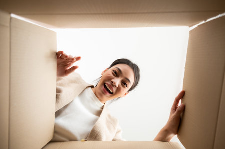 Surprised young asian woman unpacking. Opening carton box and looking inside. Packaging box, delivery service. Human emotions and facial expressionの写真素材