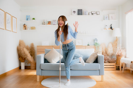 House cleaning with fun. Happy young asian housewife singing her favorite song during cleanup, using mop as microphone, enjoying domestic work. Young woman dancing and cleaning in living roomの写真素材