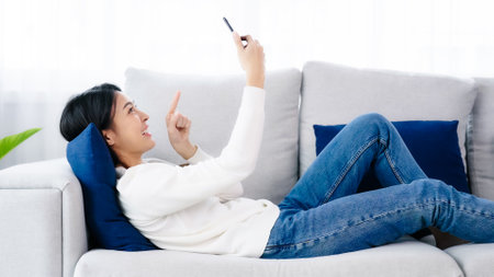 Happy young asian woman relaxing at home. Female smile lying on sofa and holding mobile smartphone. Girl using video call to friendの写真素材