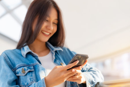 Close up hand of young asian woman smiling using mobile smart phone outdoor. Happy female tourist wearing jeans jacket and holding smartphone at publicの写真素材