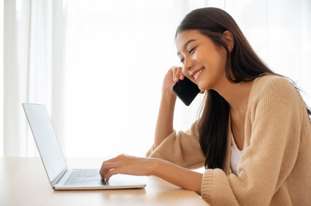 Young asian woman talking mobile smartphone while working on computer laptop on desk in living room at homeの写真素材