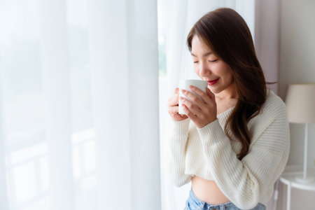 Young asian woman standing beside window and holding mug in bedroom at home, She drinking milk after wake up in the morningの写真素材