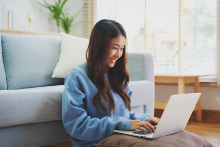 Young asian woman smile working in living room at home. Happy female using computer laptop and sitting on the floor beside couch at house, Video call with friendの写真素材
