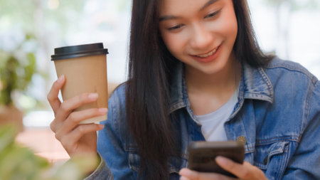 Asian woman happy and typing messages on smartphone in cafe. Smiling young woman sitting at table with hot cup of coffee and using mobile phoneの写真素材