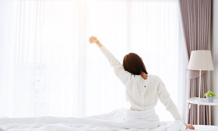 Young Asian Woman stretching in bed after waking up, back view, happy female and relaxed after good night sleep. Sweet dreams, good morning, new day, weekend, holiday, cozy and comfortの写真素材