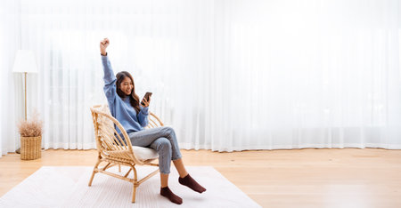 Panorama View Shot of Young Asian Woman Relaxing in Modern Wicker Chair Near Window with Sheer Curtain, Happy Female Using Mobile Smartphone Enjoying at Home, Comfort and Cozyの写真素材