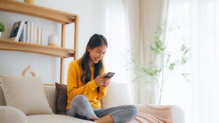 Happy young asian woman relax on comfortable couch at home, typing chat message on smartphone, smiling girl use cell phone chatting, searching information on browser wireless internet, online shoppingの写真素材
