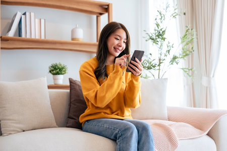 Happy young asian woman relax on comfortable couch at home texting messaging on smartphone, smiling girl use cellphone chatting, browse wireless internet on gadget, shopping online from homeの写真素材