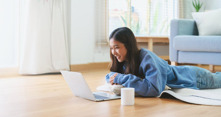 Young asian woman smile working in living room at home. Happy female using computer laptop and lying on the floor at house, Video call with friendの写真素材