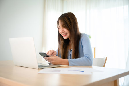 Young asian woman working on computer laptop and hand typing chat on mobile smartphone at homeの写真素材