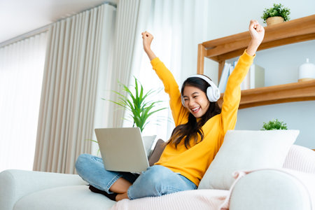 Young happy lucky woman student feeling excited winning, using computer laptop and sitting on sofa, adorable Asian female receiving great news on notebook, getting new job celebrating successの写真素材