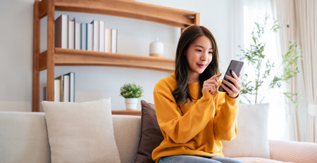 Happy young asian woman relax on comfortable couch at home texting messaging on smartphone, smiling girl use cellphone chatting, browse wireless internet on gadget, shopping online from homeの写真素材