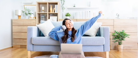 Young asian woman in good spirits working on laptop at home while sitting on the floor close to the couch. Excited female using computer notebook, wearing headphones and celebrating successの写真素材