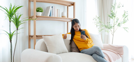 Happy young Asian woman sitting on sofa holding mobile phone using cellphone technology doing ecommerce shopping, buying online, texting messages relaxing on couch in cozy living room at homeの写真素材