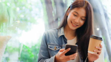 Asian woman happy and typing messages on smartphone in cafe. Smiling young woman sitting at table with hot cup of coffee and using mobile phoneの写真素材