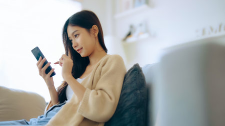 Happy young asian woman relax on comfortable couch at home texting messaging on smartphone, smiling girl use cellphone, chatting online message, shopping online from homeの写真素材