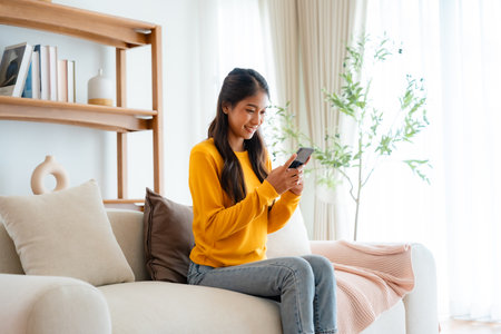 Happy young asian woman relax on comfortable couch at home texting messaging on smartphone, smiling girl use cellphone chatting, browse wireless internet on gadget, shopping online from homeの写真素材