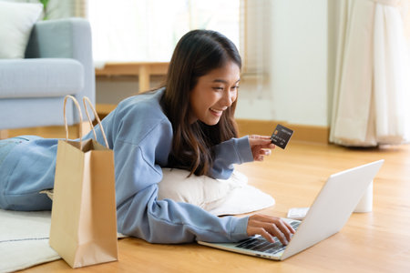 Young asian happy woman smile in living room at home, Using computer laptop, lying on the floor at house, online banking payment through the internet from bank card. Shopping online with credit cardの写真素材