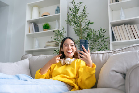 Happy young asian woman  wearing headset and relaxing on comfortable couch at home, texting messaging on smartphone, smiling girl use cellphone chatting, online shopping at home, video callの写真素材