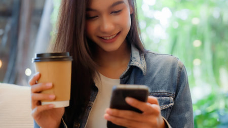 Asian woman happy and typing messages on smartphone in cafe. Smiling young woman sitting at table with hot cup of coffee and using mobile phoneの写真素材