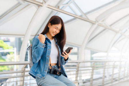Young asian woman using mobile in city. Happy female tourist wearing jeans jacket and holding smartphone at publicの写真素材