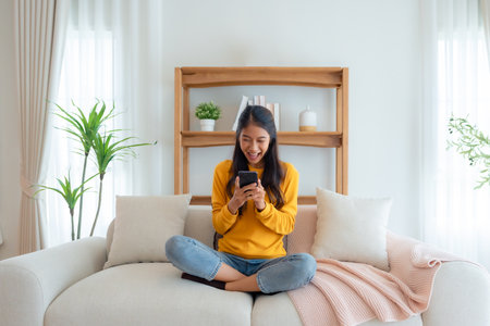 Happy young asian woman relax on comfortable couch at home texting messaging on smartphone, smiling girl use cellphone, chatting online message, shopping online from homeの写真素材