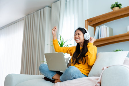 Young happy lucky woman student feeling excited winning, using computer laptop and sitting on sofa, adorable Asian female receiving great news on notebook, getting new job celebrating successの写真素材