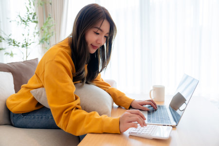 Young Asian female wearing glasses using laptop, working at home in living room, coffee mug on table. Cozy office workplace, remote workの写真素材