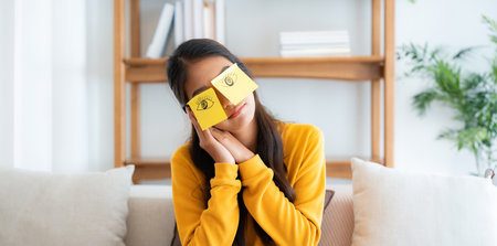 Tired adorable asian female student sleeping with stickers on eyes, sitting on couch. Beautiful...