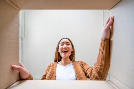 Low angle view of surprised young asian woman unpacking. Opening carton box and looking inside....