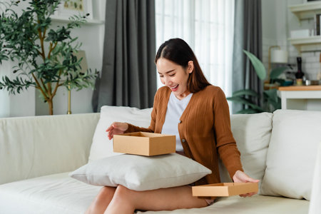 Happy young Asian woman opening delivery package at home. Beautiful female customer open box and sitting on couch in living roomの写真素材