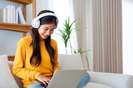 Young adorable asian woman smile wearing headset working in living room at home. Happy gorgeous female using computer laptop and sitting on couch at house, Video call with friendの写真素材