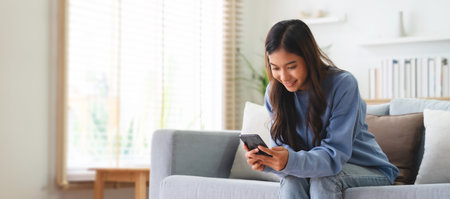 Happy young asian woman relax on comfortable couch at home, typing chat message on smartphone, smiling girl use cell phone chatting, searching information on browser wireless internet, online shoppingの写真素材
