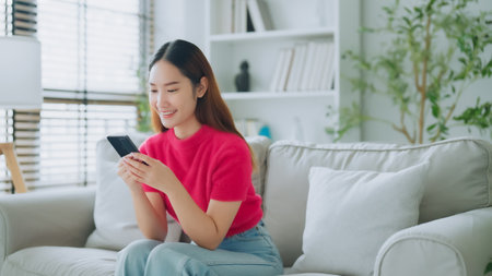 Happy young asian woman relax on comfortable couch at home texting messaging on smartphone, smiling girl use cellphone chatting, online shopping at home, video call communicationの写真素材