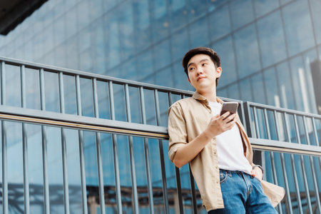 Portrait of handsome Asian student using smartphone. A young man standing outdoor happy smiling with holding mobile phoneの写真素材