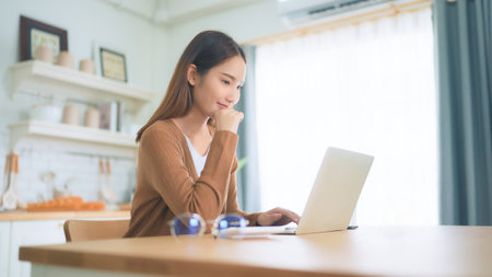 Beautiful young Asian woman working with laptop computer in home kitchen. Work at homeの写真素材