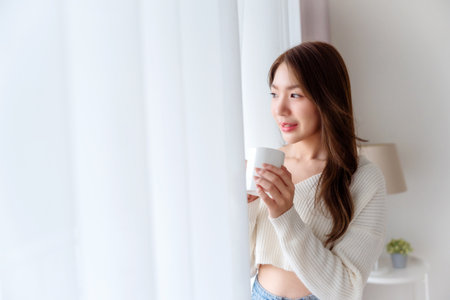 Young asian woman standing beside window and holding mug in bedroom at home, She drinking milk after wake up in the morningの写真素材