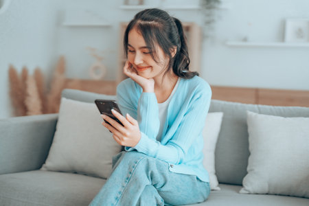 Smiling pretty young Asian woman sitting on cozy couch, using funny mobile apps in living room. Woman at Home, Doing Online Shopping, Messaging Friends, Posting on Social Mediaの写真素材