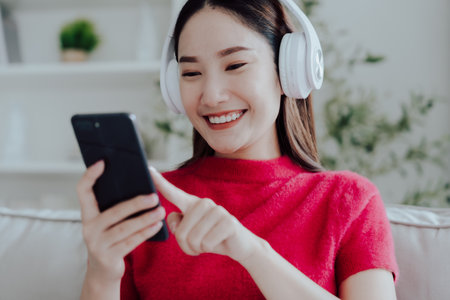 Young Asian woman relaxing on sofa at home, listening music with headset on cell phone. People lifestyle conceptの写真素材