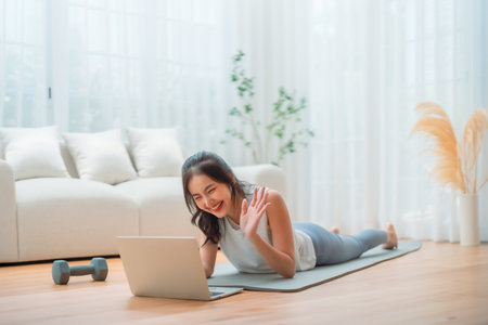 Young Asian woman lying on the floor on yoga mat in front of laptop and training at home. Beautiful smiling woman doing stretching exercise while communicates with a trainer online before workoutの写真素材