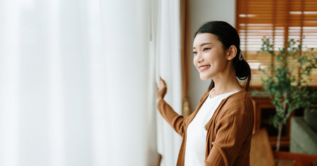 Happy Asian woman standing by the window and looking outside the living room enjoying the bright sunshineの写真素材