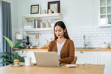 Beautiful young Asian woman working with laptop computer in home kitchen. Work at homeの写真素材
