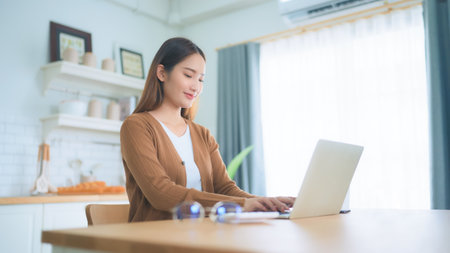 Beautiful young Asian woman working with laptop computer in home kitchen. Work at homeの写真素材