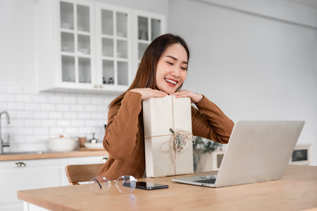 Young asian woman using computer laptop at home. Female showing gift box while on video conversation with friend. Happy birthday, Happy new year, Thanksgivingの写真素材