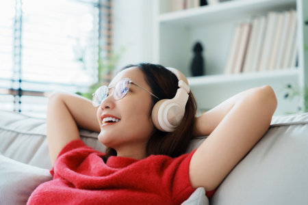 Relaxed young Asian woman enjoying rest on comfortable sofa, calm attractive girl relaxing on couch, breathing fresh air, wearing eyeglasses and headset, meditating at homeの写真素材