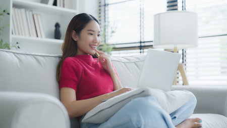 Happy young Asian woman using laptop while seated on couch at homeの写真素材
