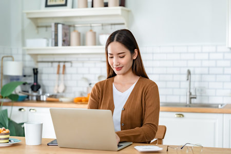 Beautiful young Asian woman working with laptop computer in home kitchen. Work at homeの写真素材