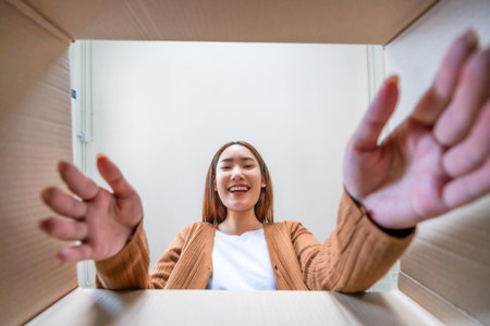 Low angle view of surprised young asian woman unpacking. Opening carton box and looking inside. Packaging box, delivery service. Human emotions and facial expressionの写真素材