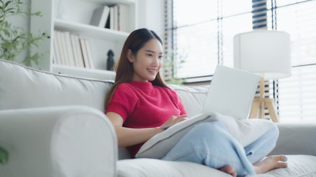 Happy young Asian woman using laptop while seated on couch at homeの写真素材
