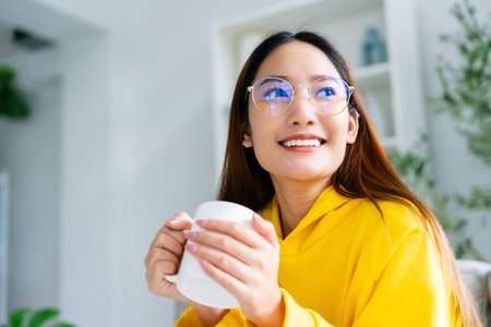 Young asian woman wearing eyeglass sitting on sofa and holding mug in living room at home, drinking milk or tea in the morning. Healthy and refresh moodの写真素材